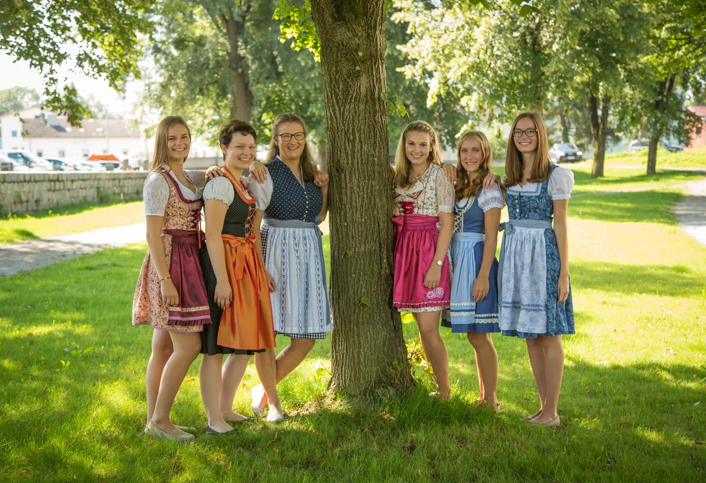 Sechs junge Frauen in Dirndln lachen gemeinsam an einem Baum im Sommerlicht