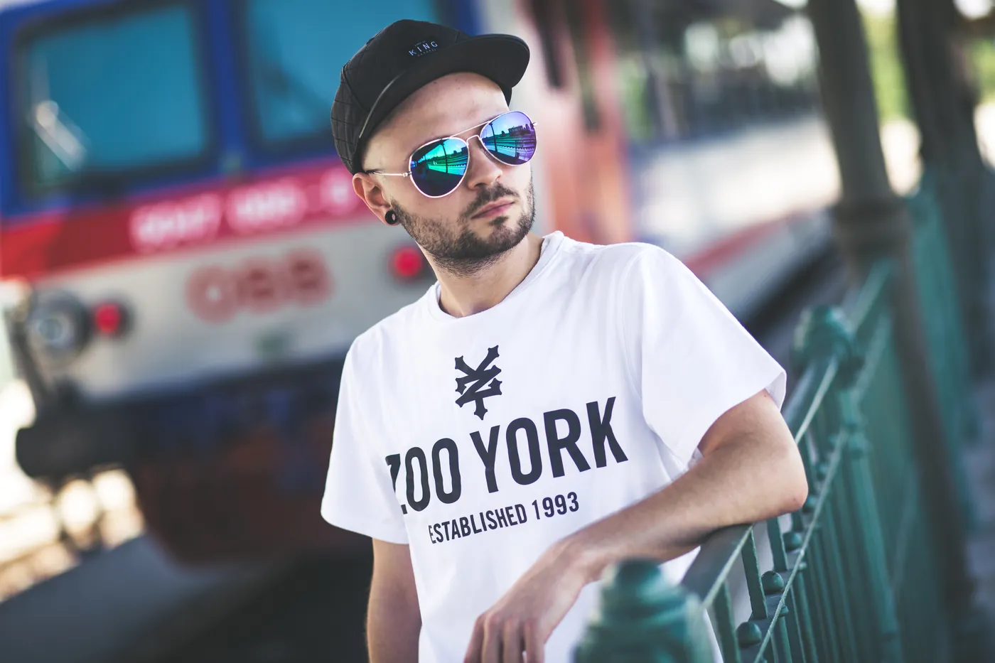 Mann mit Kappe und verspiegelter Sonnenbrille am Geländer, im Hintergrund ein Zug
