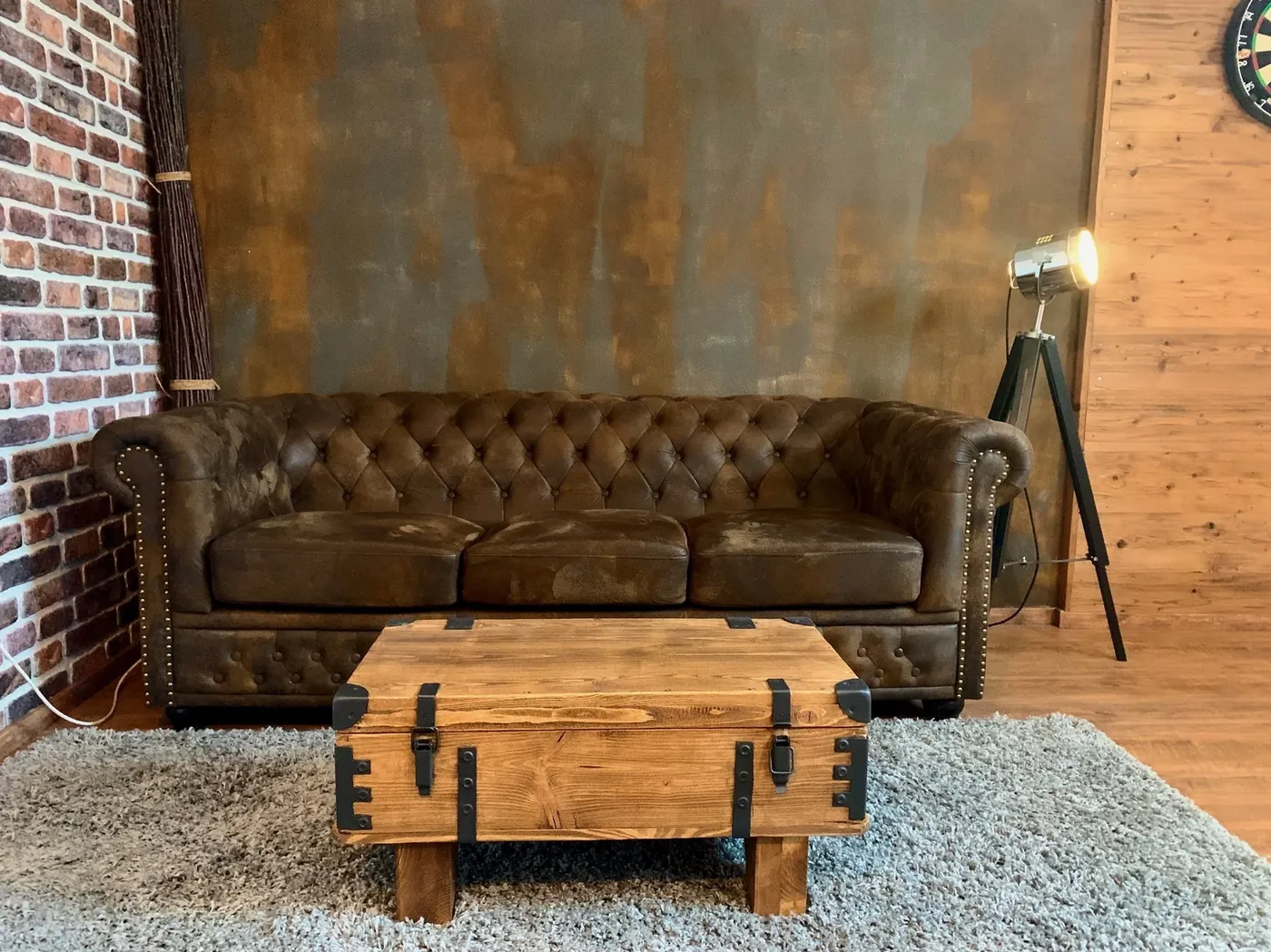 Studioecke mit braunem Chesterfield-Ledersofa vor fleckig gemalter Hintergrundwand, davor eine Holztruhe als Tisch auf grauem Hochflorteppich, rechts eine Stativ-Leuchte